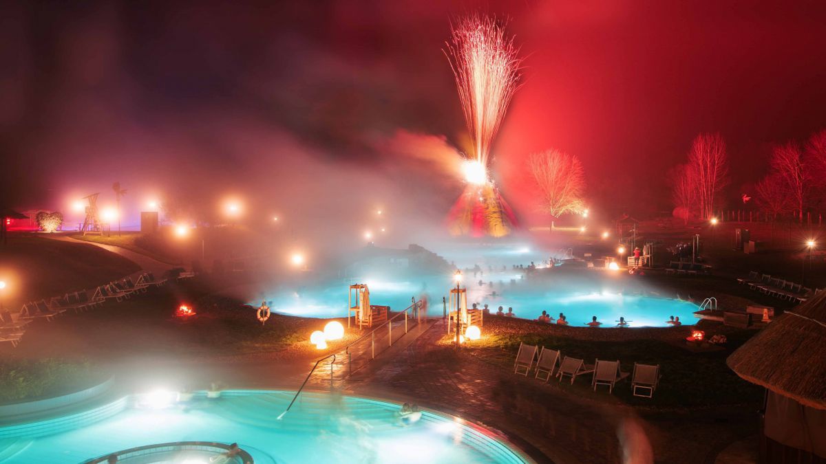 Vulkania Thermenlandschaft bei Nacht. Mehrere Becken, dahinter eine Lichtshow - der Vulkanhügel speit rote Strahlen in den Nachthimmel.