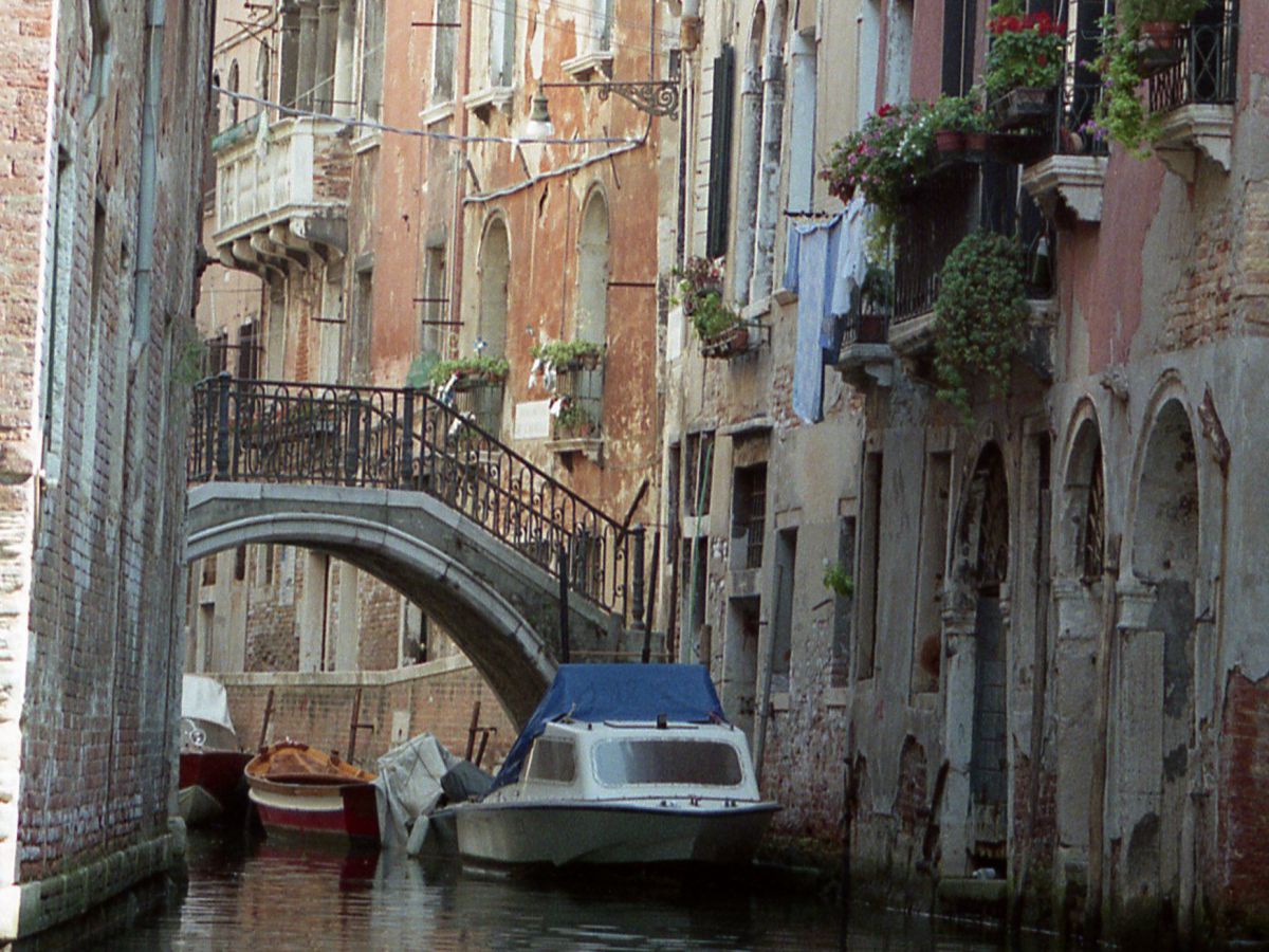 Enger Seitenkanal in Venedig, zwischen den Häusern eine Brücke, einige Boote entlang der Hauswand..