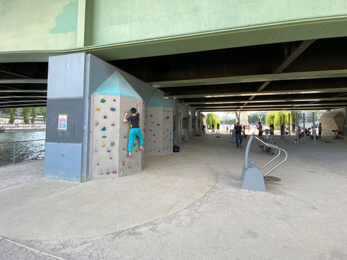 Unter einer Brücke sind Sportgeräte aufgebaut, eine Frau klettert an einer Kletterwand am Brückenpfeiler.