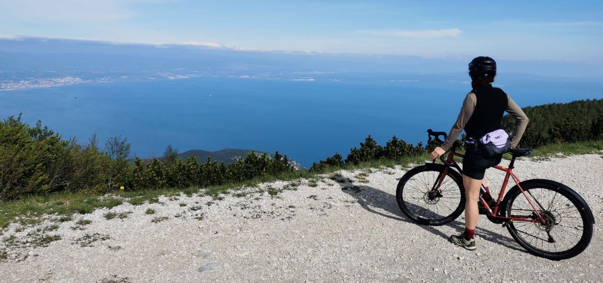 Radfahrer steht auf einer Anhöhe auf einem Schotterweg und blickt hinunter in eine Meeresbucht.