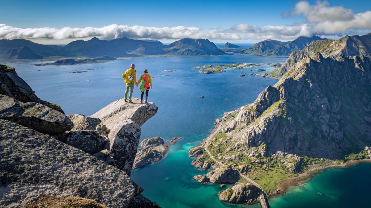 Zwei Personen aug einem Felsvorsprung hoch über einer Fjordlandschaft.