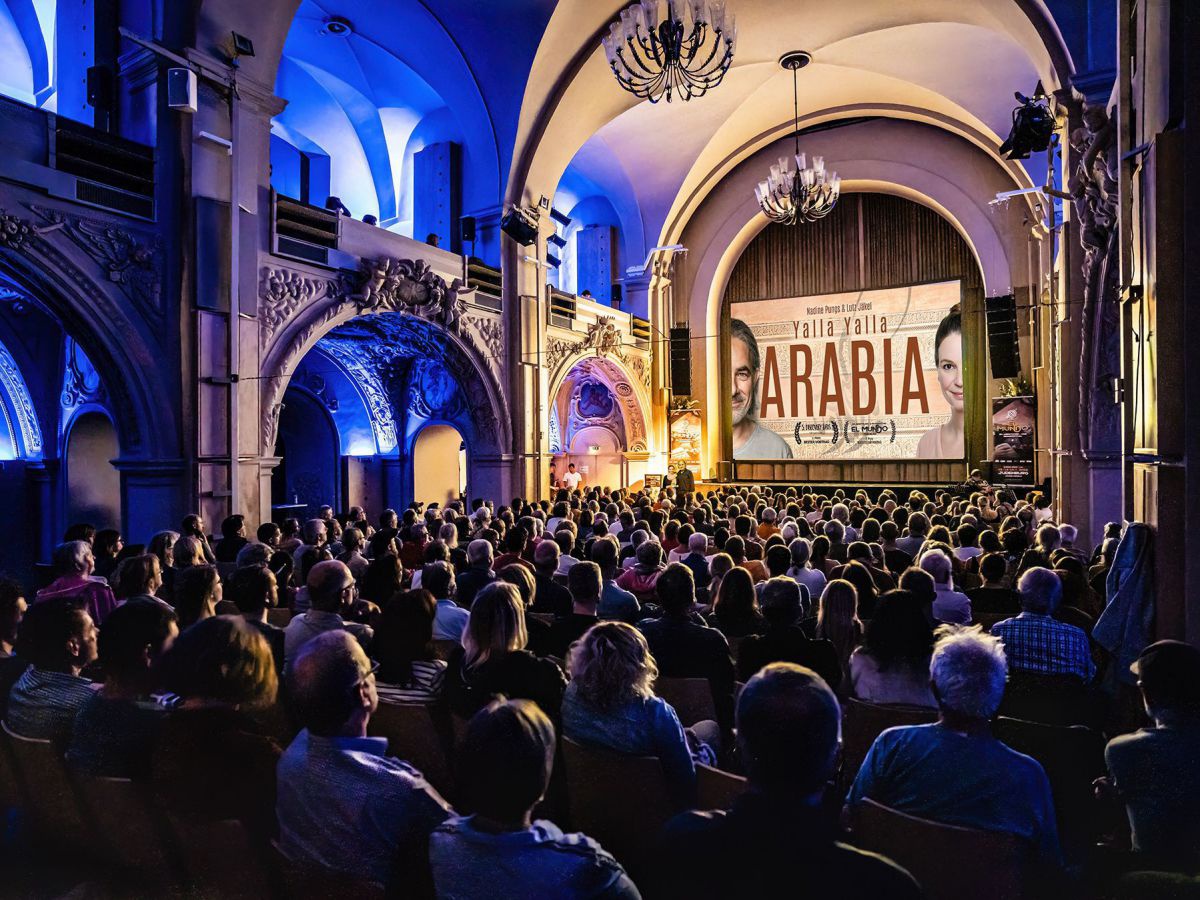 Abgedunkelter Festsaal. Menschen blicken zur Bühne, wo ein großes Poster mit dem Schriftzug ARABIA zu sehen ist.