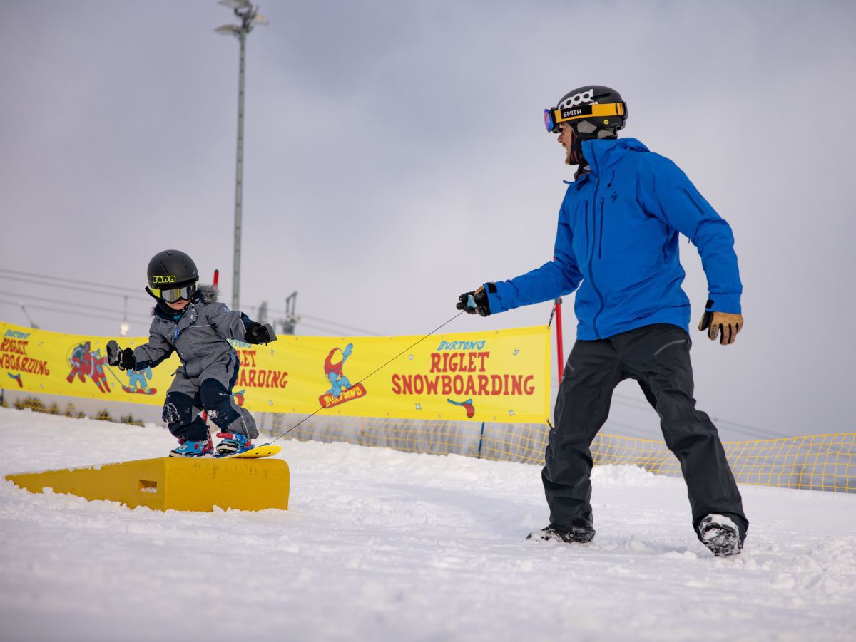 Kind fährt mit Snowboard über eine Schanze, Mann unterstützt es dabei.