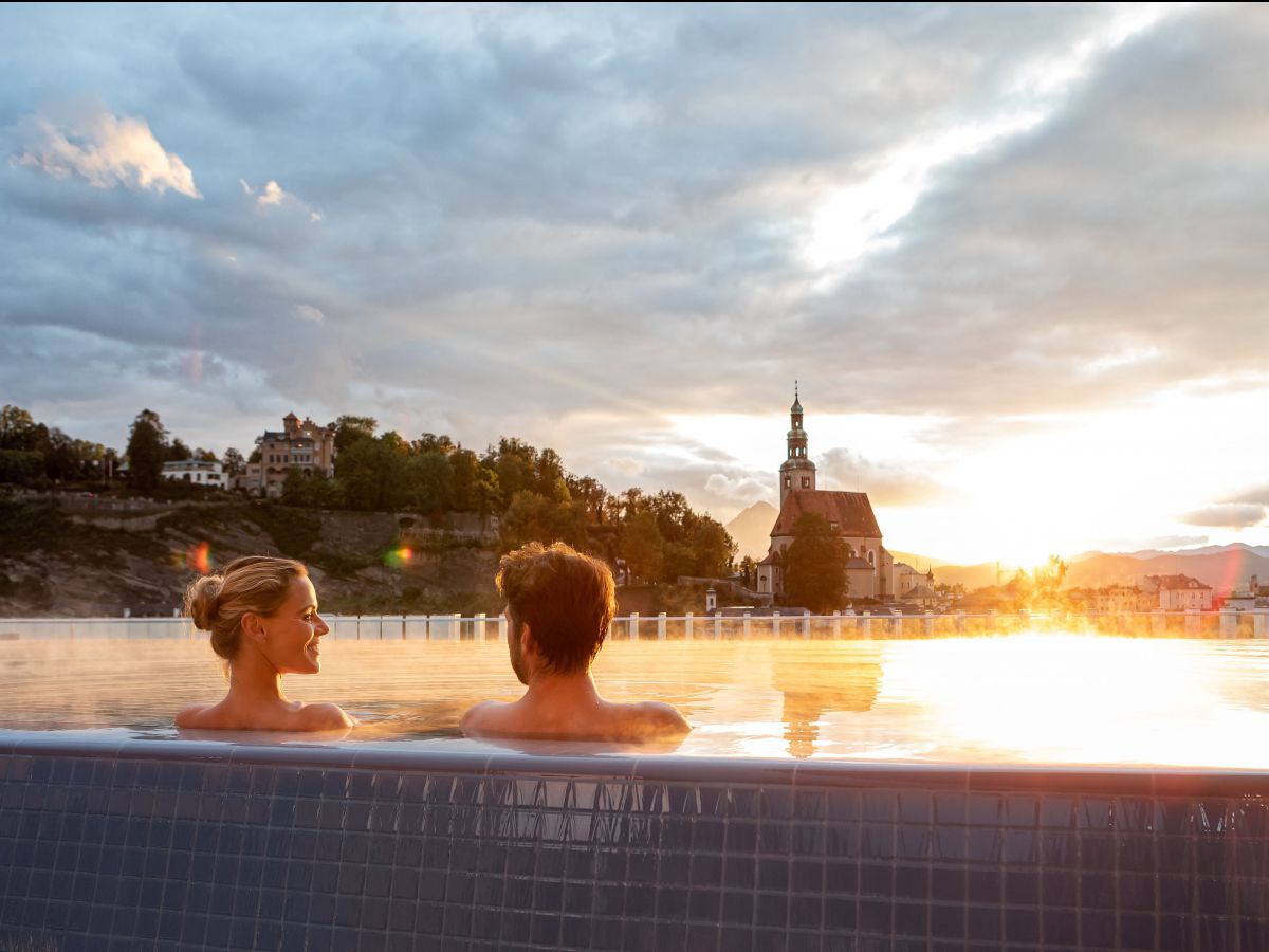 Mann und Frau sitzen im Infinitypool bei Sonnenuntergang und blicken auf eine Kirche.