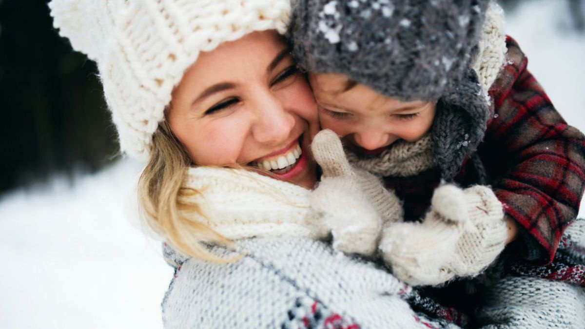 Eine junge Frau mit Kleinkind auf dem Arm, beide wollig-warm eingepackt in Jacken, Hauben und Fäustlingen. Im Hintergrund Schneelandschaft. Beide lachen ausgelassen.