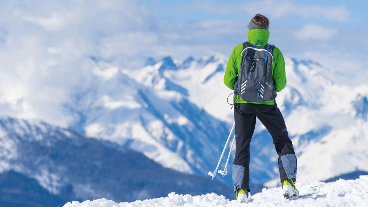 Eine Person in Winterkleidung und Schiausrüstung steht mit dem Rücken zum Betrachter auf einem Berg im Schnee und blickt auf das weite, winterlich weiße Gipfelpanorama.