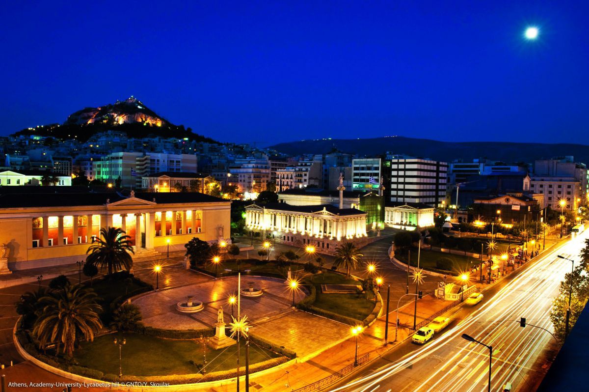 Ein festlich beleuchteter Platz in Athen bei Nacht. Gebäude mit Säulen, Grünflächen, Palmen und Springbrunnen. Der Himmel ist in tiefes dunkelblau getaucht, rechts im oberen Eck sieht man einen vollen Mond. Am Horizont ein bewaldeter Hügel, ebenfalls mit 