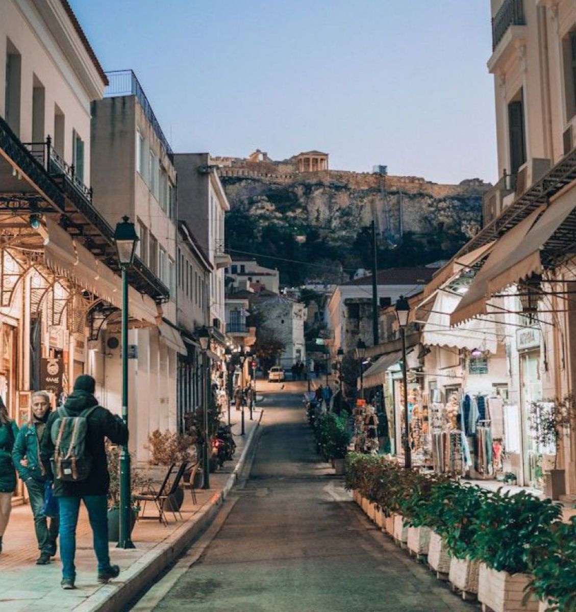 Einige wenige warm bekleidete Fußgänger in einer Einkaufsstraße in Athen, oben am Hügel sieht man im Hintergrund die Akropolis.