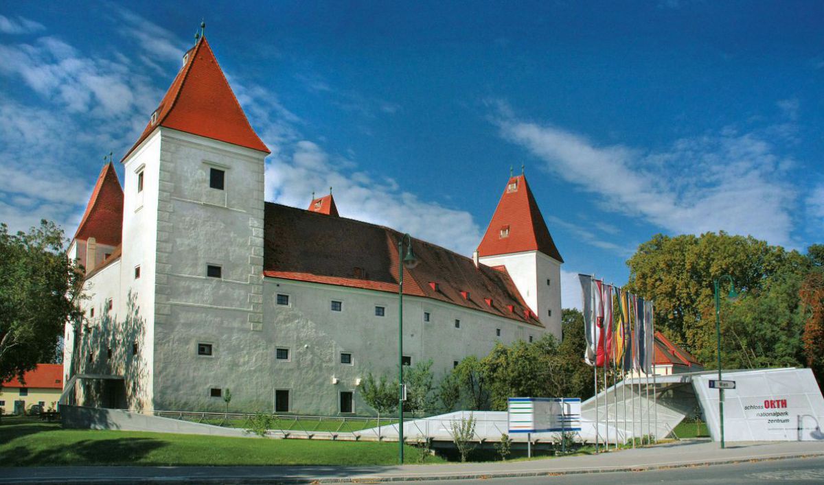 Außenansicht vom Schloss Orth. Mächtiger dreigeschoßiger weißer Bau mit steilem roten Dach, an den Ecken jeweils ein etwas höherer Turm. Vor dem Eingang mehrere Fahnenstangen und Fahnen.