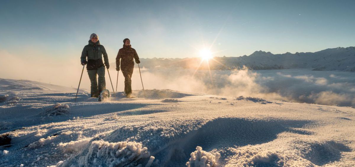 Paar mit Schneeschuhen unterwegs auf einer Hochebene, im Hintergrund geh1 die Sonne über den Bergen auf.