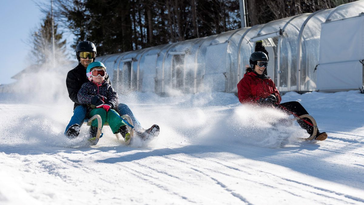 2 Rodeln sausen die Piste runter, auf einer ein Mann mit einem Kind, auf der anderen ein Kind alleine.