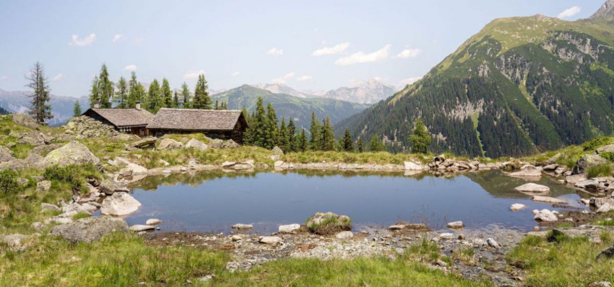 Kleiner Bergsee, dahinter lugen 2 Hüttendächer hervor, Bergpanorama.