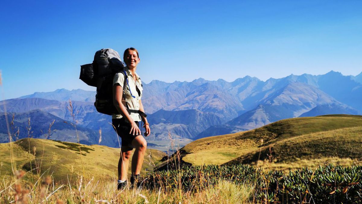 Andrea Ensmann am Omalo-Trail in Georgien: Die Frau steht in kurzärmeliger Wanderkleidung und schwer bepackt mit einem großen Rucksack auf einem sanft hügeligenm grün bewachsenen Höhenwanderweg. Im Hintergrund schimmert bläulich ein zerklüftetes Gebirgsma