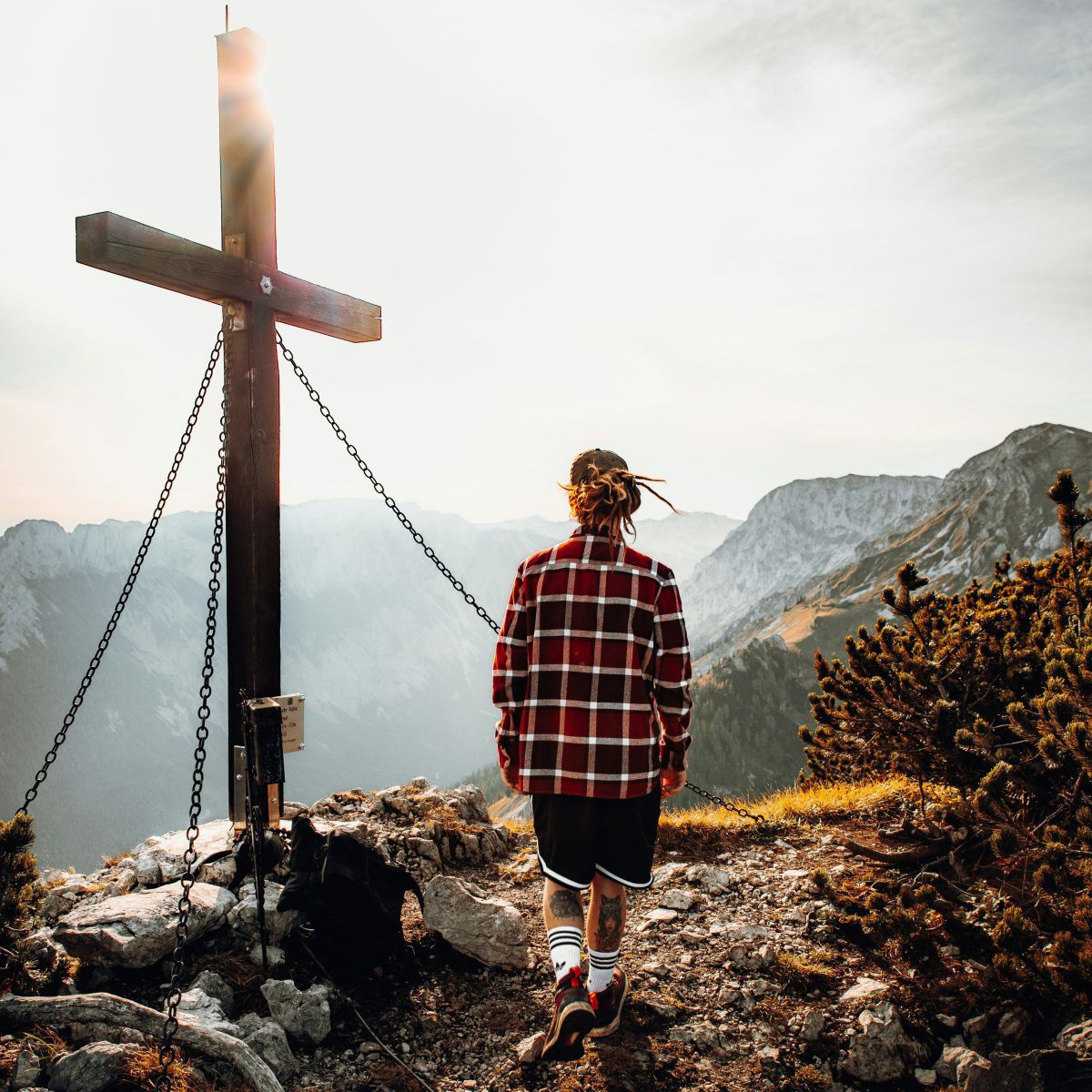 Eine Person in sportlichen schwarzen Shorts und kariertem Hemd geht auf ein von der Sonne beleuchtetes Gipfelkreuz zu. Im Hintergrund noch mehr Bergpanorama.