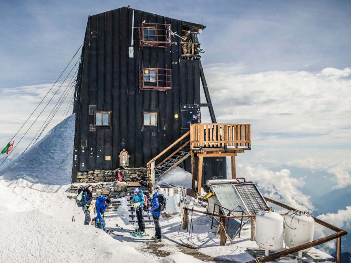 Auf einem schneebedeckten Gipfel steht, mit Seilen gesichert, eine hohe, schmale Hütte mit schwarzer Holzverkleidung und wenigen kleinen Fenstern. Rundum sieht man hauptsächlich Wolken und einige tiefergelegene Berggipfel.