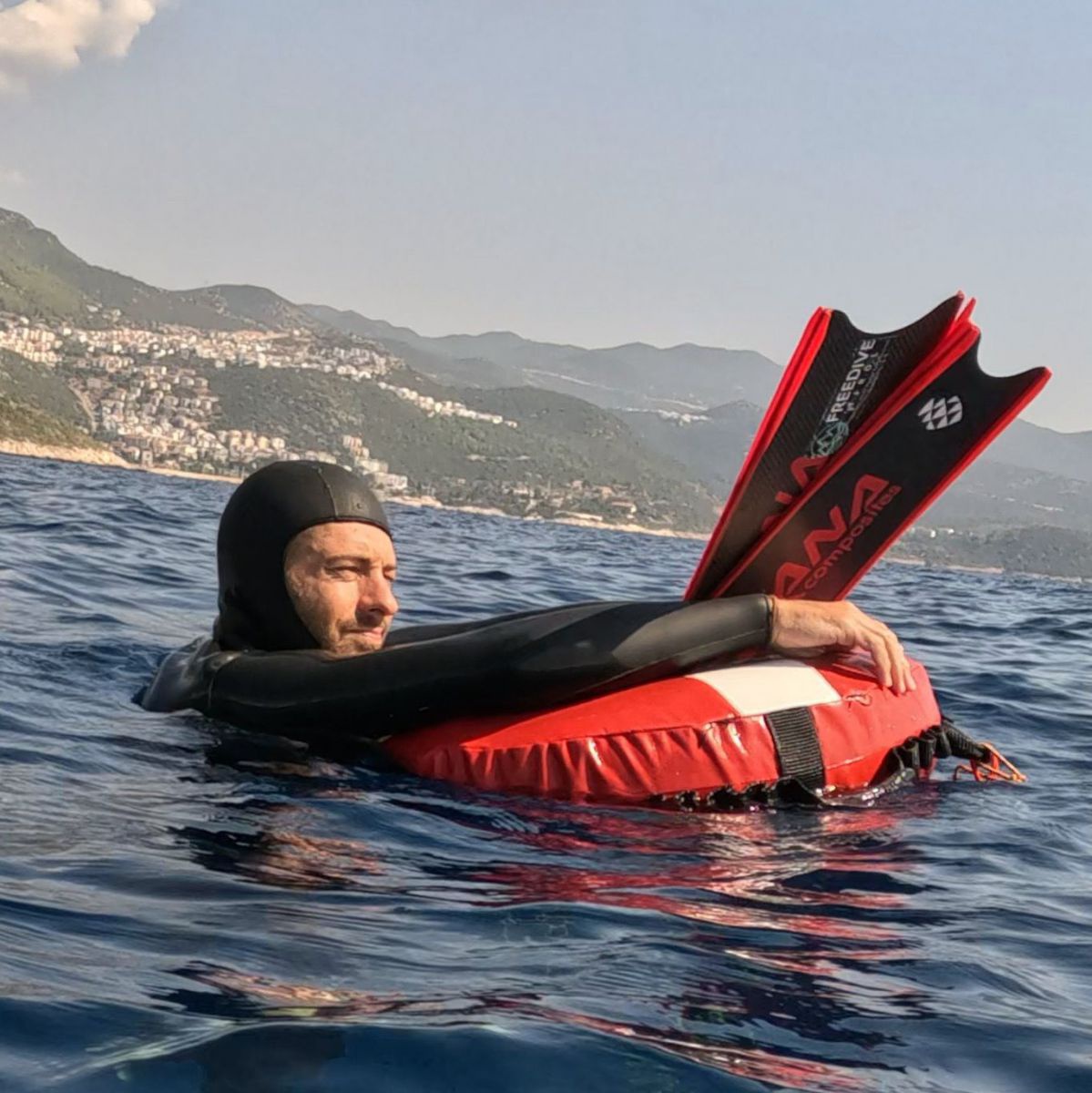 Kai Kipfmüller im Ganzkörper-Neoprenanzug mit Kapuze ruht sich nach einem Tauchgang auf einem im See treibenden Schwimmreifen aus. Die Flossen hält er in den Händen.