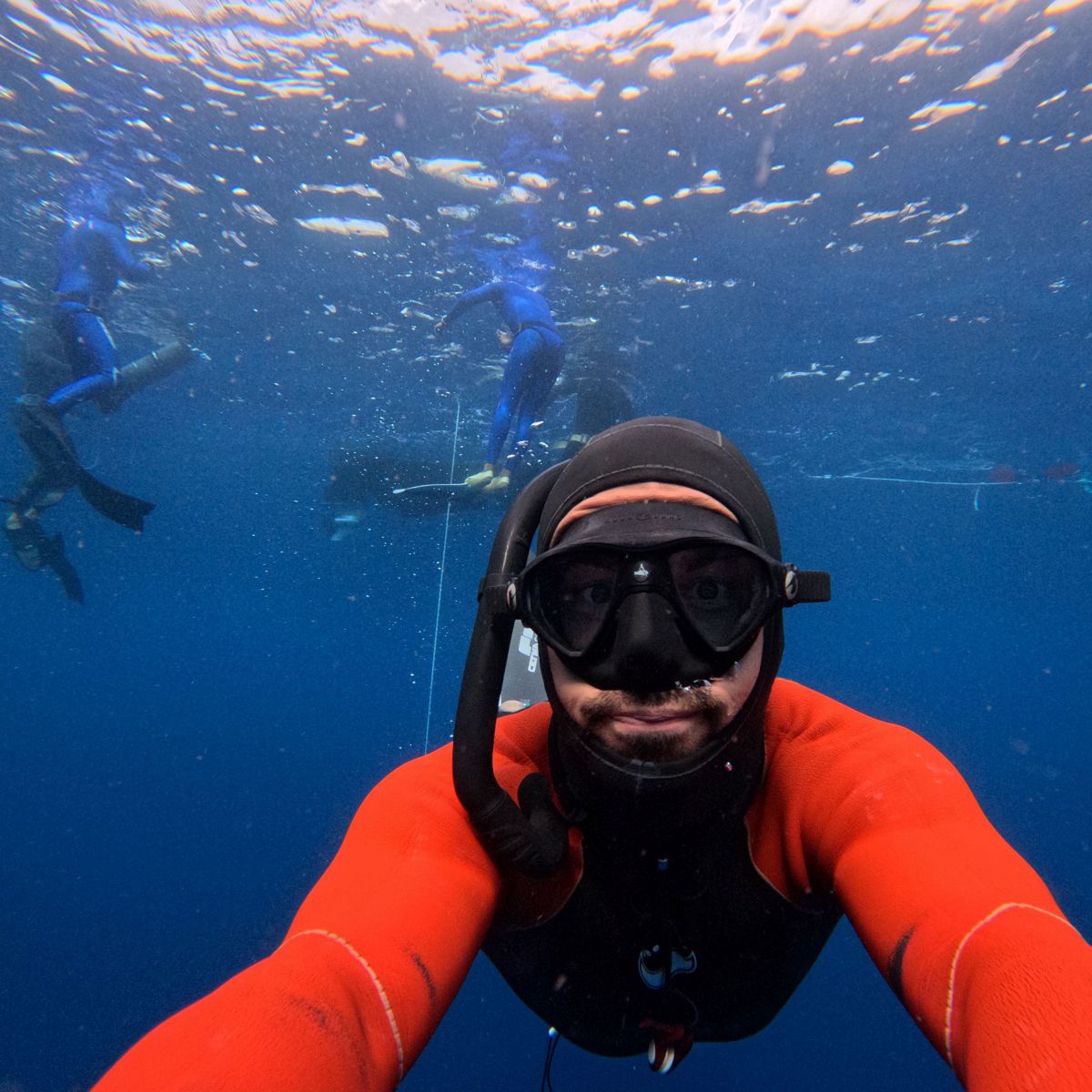 Selfie eines Tauchers unter Wasser. Er trägt Neopren Anzug, Taucherbrille und Schnorchel. Im Hintergrund sieht man weitere Taucher an der Oberfläche des tiefblauen Sees. 