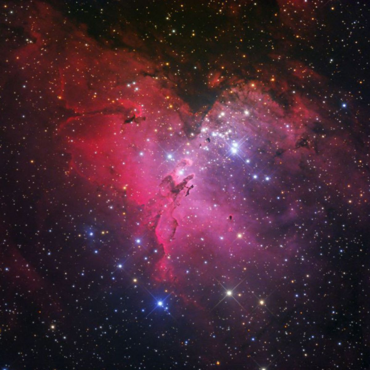 Foto eines Sternhaufens umgeben von Nebel. Auf schwarzem Hintergrund strahlen unzählige verschieden große Lichtpunkte in bläulich-weißem Licht. Der Nebel leuchtet in Rot- über Rosa- bis Lila- und Blautönen.