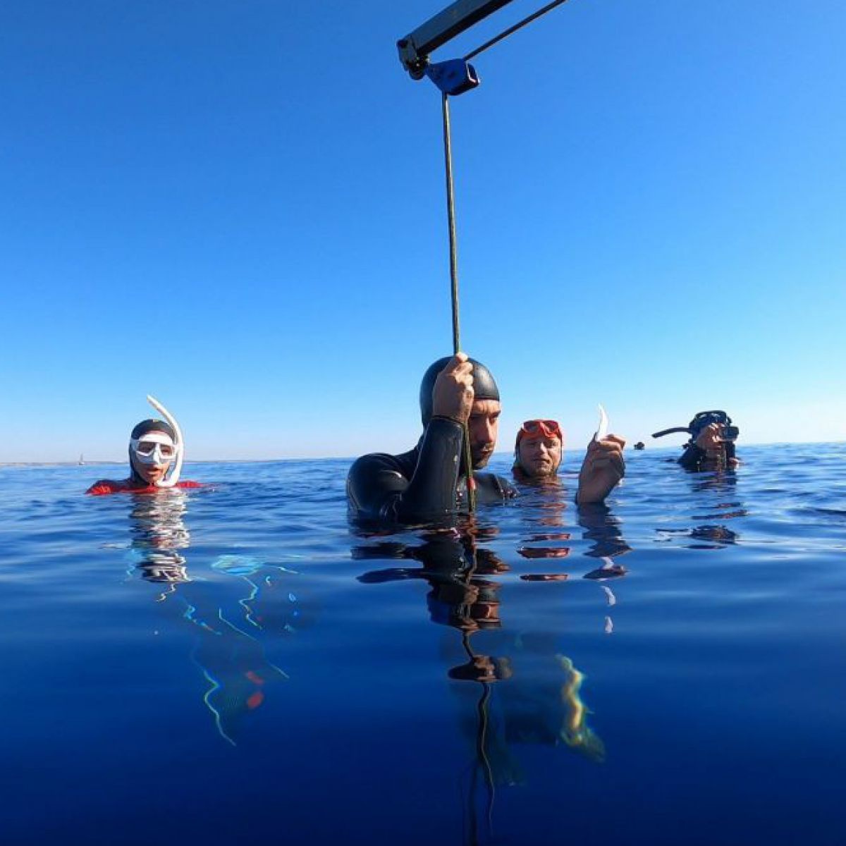 Eine Gruppe von Personen in Neoprenanzügen und teilweise mit Taucherbrillen ausgestattet halten sich an der Wasseroberfläche auf, um den Erklärungen des Trainers zu folgen. Der Trainer hält sich gerade mit einer Hand am Drahtseil eines Krans fest.