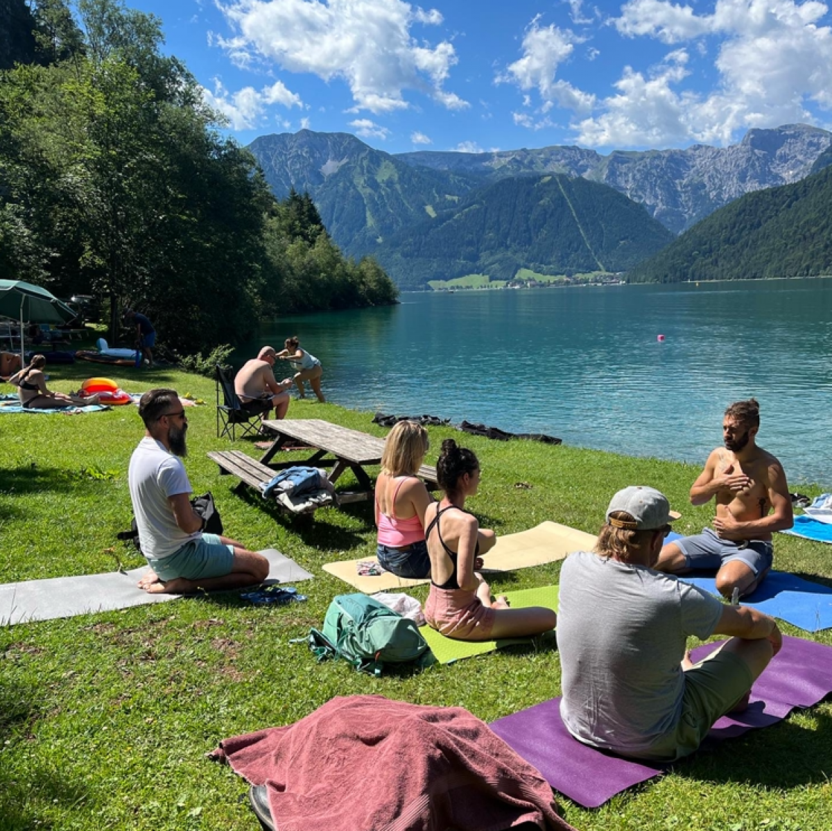 Eine Gruppe Personen sitzen auf Yogamatten auf der Wiese am Seeufer und folgen den Atemübungen des Trainers.