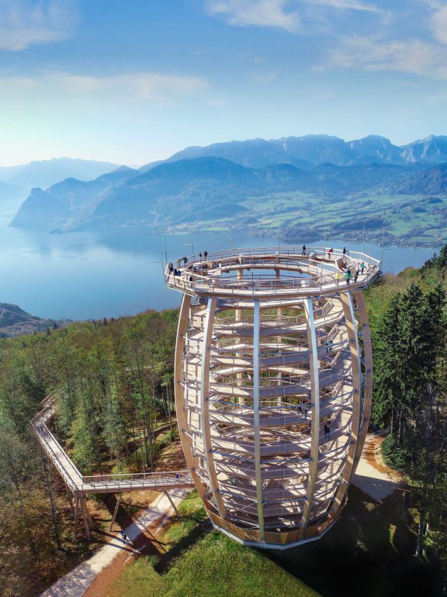 Aussichtsturm aus ellipsenförmiger Konstruktion auf einem bewaldeten Berghang. Hözerne Stege des Baumwipfelpfades führen zum Turm. Im Tal liegt ein See inmitten der Bergketten.