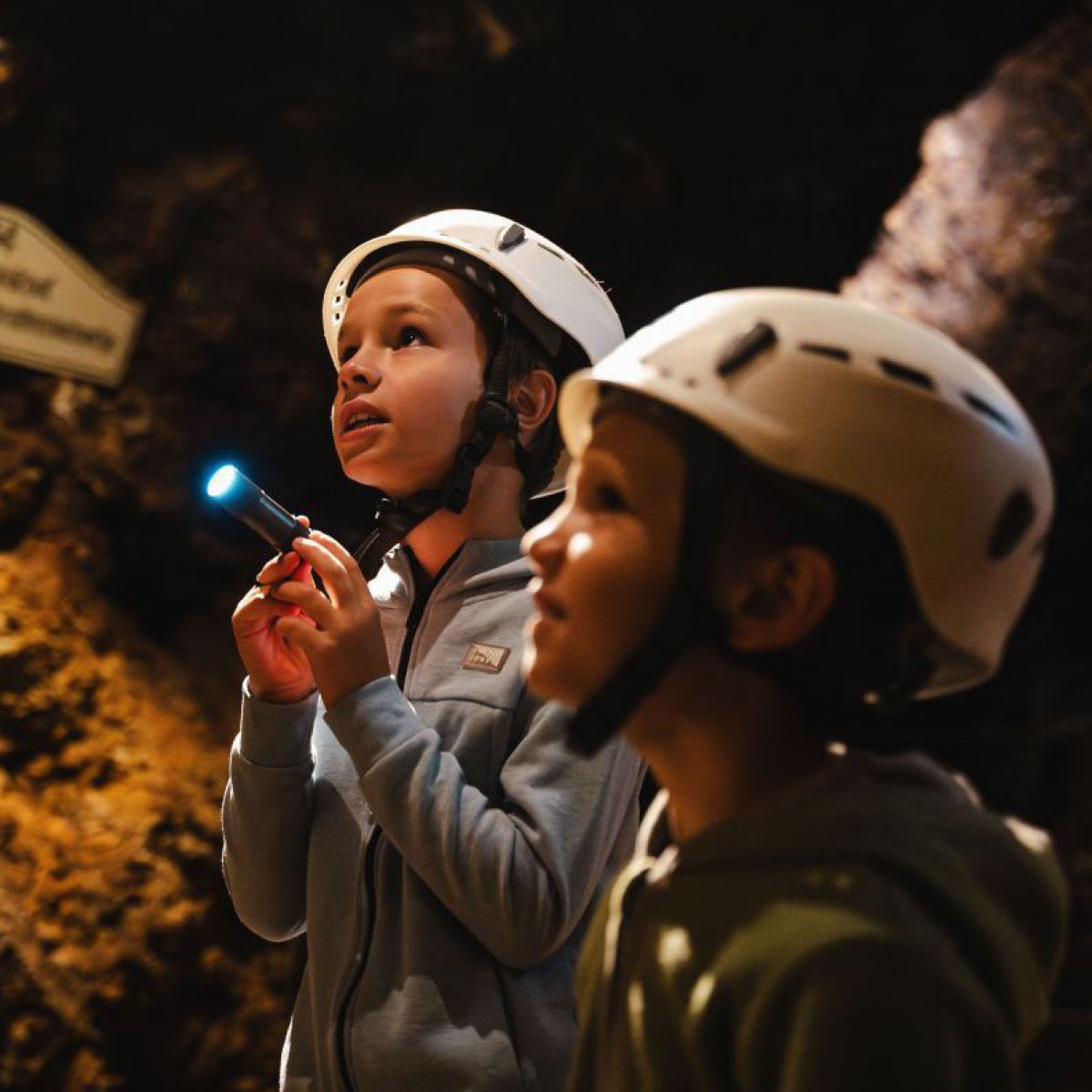 Zwei Kinder im Kindergartenalter mit weißen Schutzhelmen auf den Köpfen in einer Höhle. An der Felswand sind Schilder angebracht. Ein Kind leuchtet mit einer Taschenlampe. Beide blicken gespannt und interessiert.