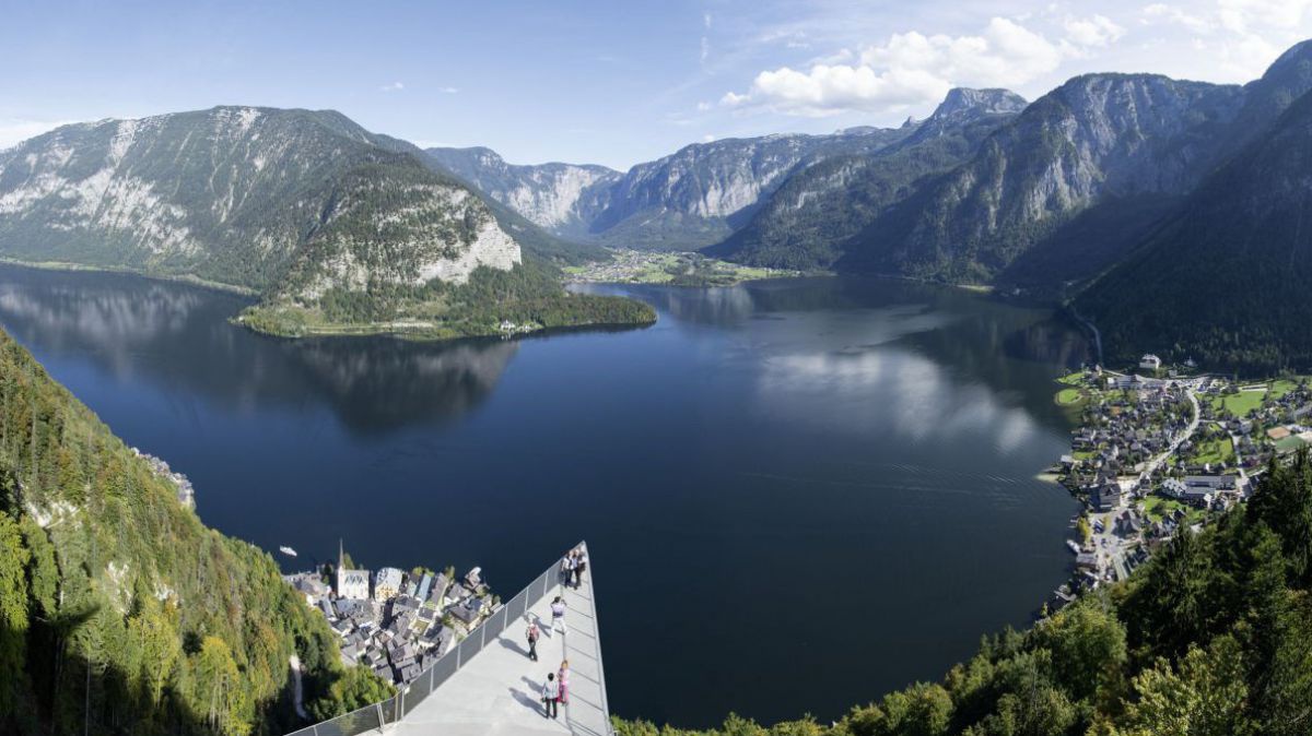 Blick aus der Höhe hinunter auf den See, der zwischen Bergketten eingebettet liegt. Im Bildvordergrund ragt die Spitze einer Aussichtsplattform hervor.