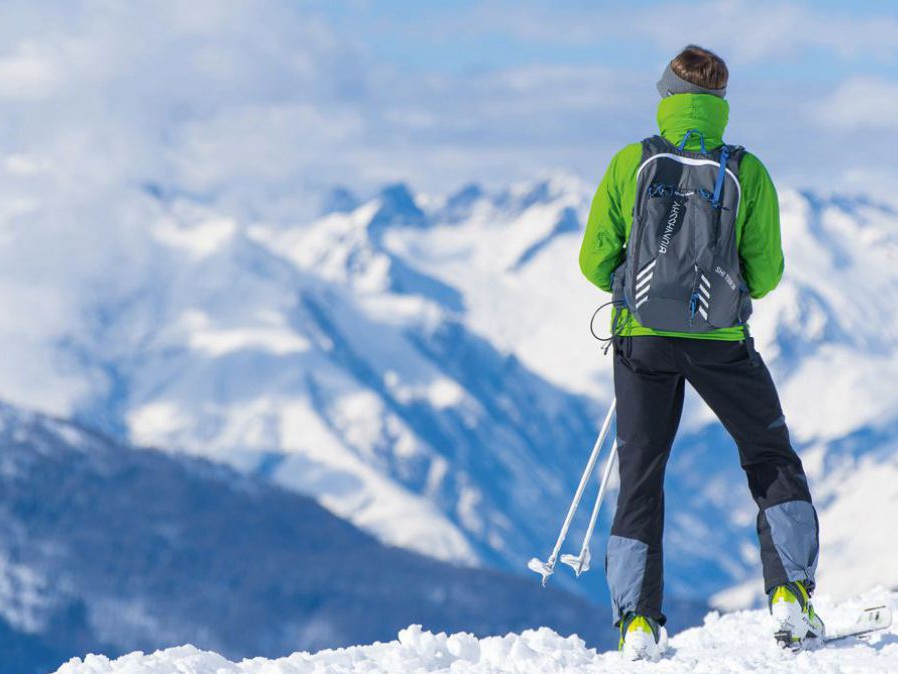 Eine Person in Winterkleidung und Schiausrüstung steht mit dem Rücken zum Betrachter auf einem Berg im Schnee und blickt auf das weite, winterlich weiße Gipfelpanorama.