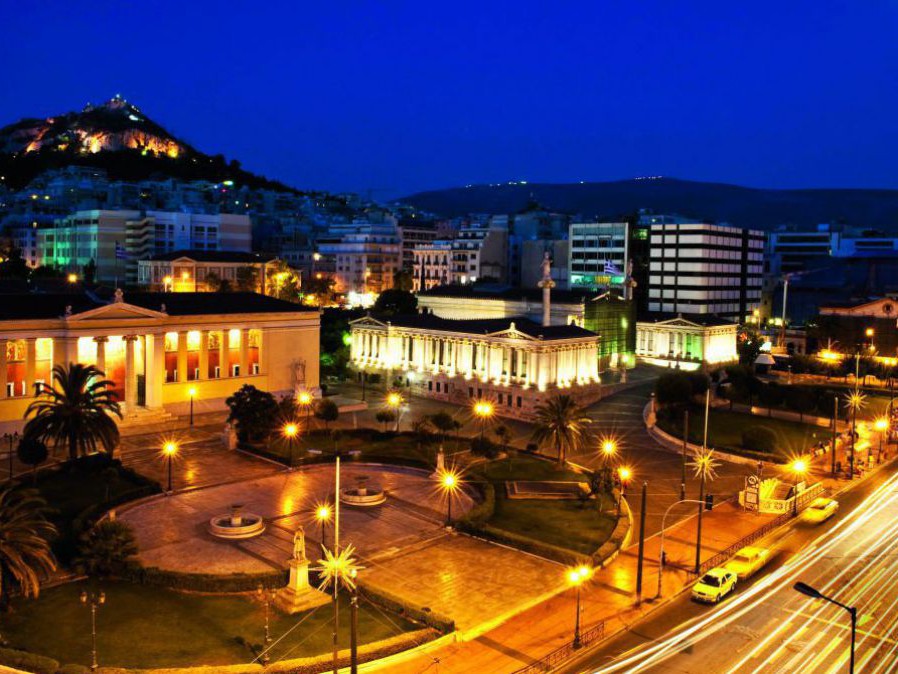 Ein festlich beleuchteter Platz in Athen bei Nacht. Gebäude mit Säulen, Grünflächen, Palmen und Springbrunnen. Der Himmel ist in tiefes dunkelblau getaucht, rechts im oberen Eck sieht man einen vollen Mond. Am Horizont ein bewaldeter Hügel, ebenfalls mit 