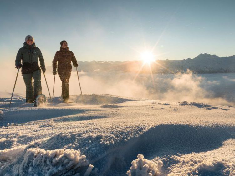 Paar mit Schneeschuhen unterwegs auf einer Hochebene, im Hintergrund geh1 die Sonne über den Bergen auf.